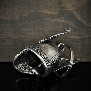 May include: Silver-colored motorcycle bell with wing accents and the word "BRAVO" engraved on the side. The bell is lying on a reflective black surface with a wooden background.
