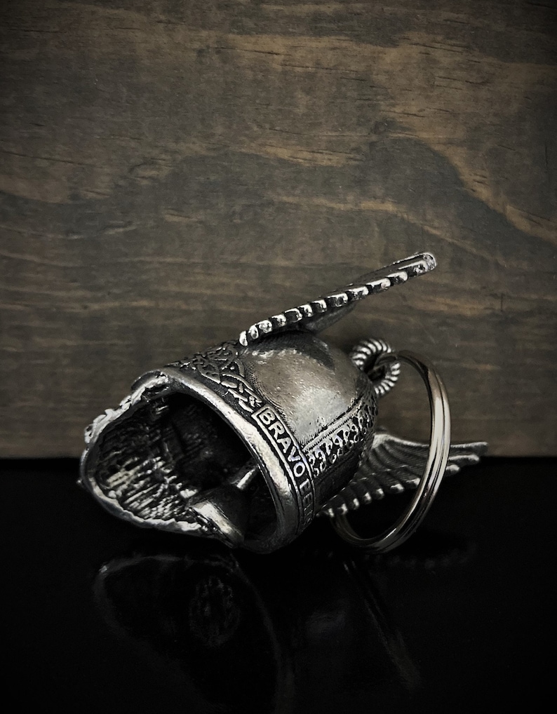 May include: Silver-colored motorcycle bell with wing accents and the word "BRAVO" engraved on the side. The bell is lying on a reflective black surface with a wooden background.