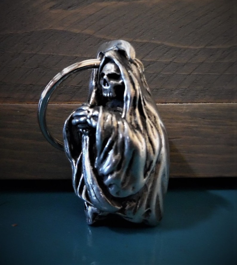 Puede incluir: Llavero de metal plateado con una escultura detallada de la Muerte, con una t&uacute;nica con capucha y sosteniendo una guada&ntilde;a. El llavero tiene un anillo circular para sujetar las llaves.