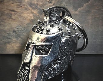 May include: A silver-toned metal keychain shaped like a detailed warrior helmet with a prominent crest, rivets, and a textured beard. The helmet features a face mask with eye openings and is attached to a circular metal split ring for keys.