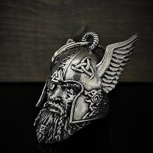 May include: A silver-colored Viking helmet keychain with intricate detailing. The helmet features a bearded face, winged accents, and Celtic knot designs. The keychain is made of metal and has a key ring.