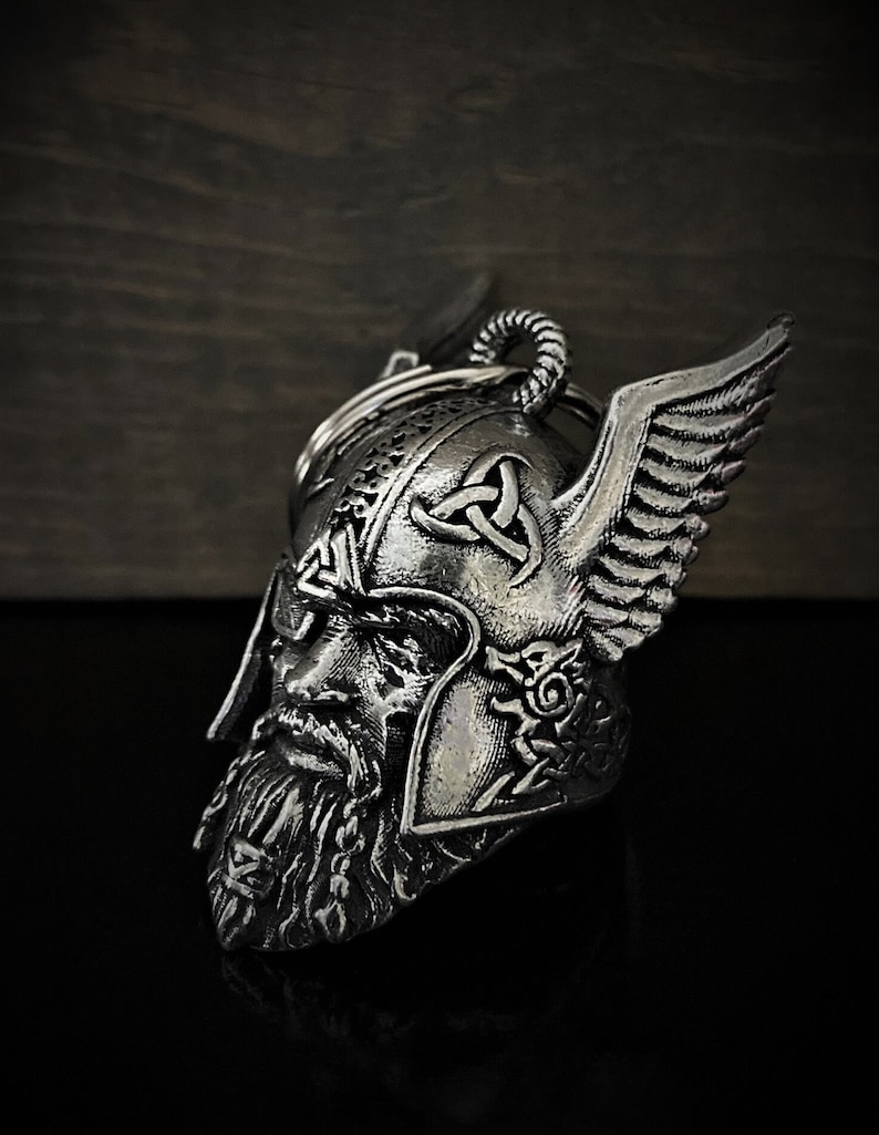 May include: A silver-colored Viking helmet keychain with intricate detailing. The helmet features a bearded face, winged accents, and Celtic knot designs. The keychain is made of metal and has a key ring.