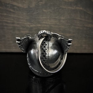 May include: A silver-colored, spherical pendant with intricate detailing and small wings on either side. A large ring is attached to the top. The pendant is set against a dark, textured background.