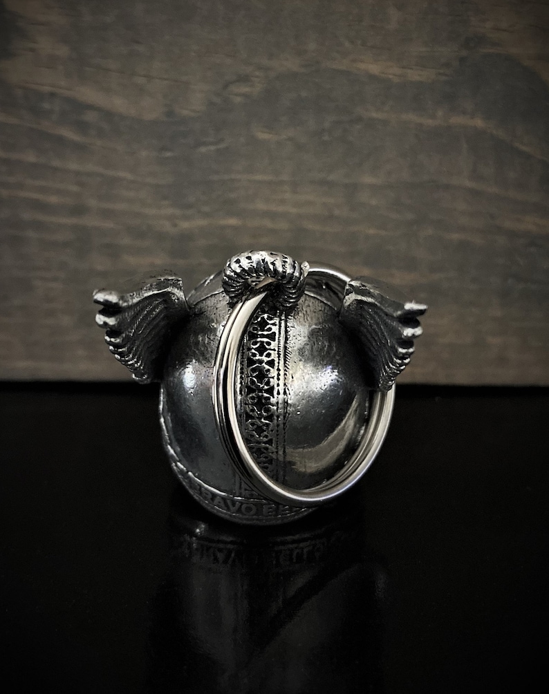May include: A silver-colored, spherical pendant with intricate detailing and small wings on either side. A large ring is attached to the top. The pendant is set against a dark, textured background.