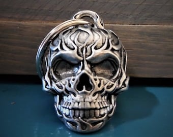 May include: Silver skull keychain with a flame design. The skull has a detailed face with a ring attached to the top.