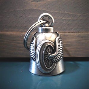 May include: A silver-toned metal bell charm featuring a raised relief design of a motorcycle wheel flanked by detailed wings. The bell has a smooth, rounded top with a sturdy metal split ring attached to the loop for secure hanging.