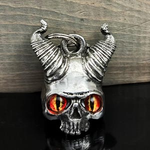 May include: A silver skull-shaped keyring with large curved horns and fiery red and yellow eyes. The skull has detailed facial features and is attached to a metal keyring. The background is a wooden surface.