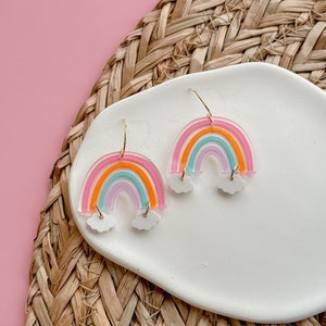 May include: A pair of rainbow-colored acrylic earrings with white cloud accents. The earrings are on a white plate with a woven background.