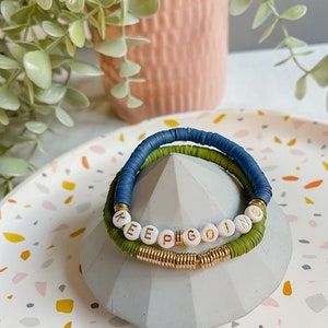 May include: Three bracelets stacked on a grey concrete base. The top bracelet is blue and green, the middle bracelet is gold, and the bottom bracelet has white beads with the words "KEEP GOING" in gold lettering.