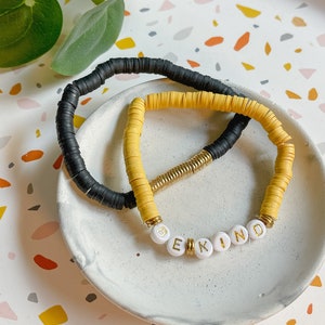 Puede incluir: Dos pulseras, una negra y otra amarilla, con la palabra "BE KIND" escrita en cuentas blancas con letras doradas.
