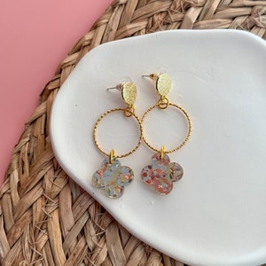 May include: Gold hoop earrings with a small gold disc at the top and a four-leaf clover charm with a glittery finish in shades of gold, silver, and pink.