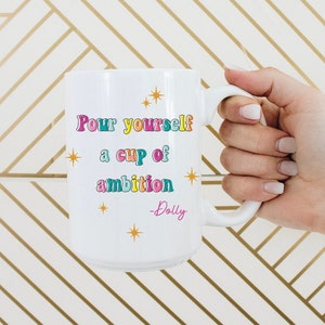 May include: White ceramic mug with a gold, pink, blue, and green design. The mug says "Pour yourself a cup of ambition - Dolly".