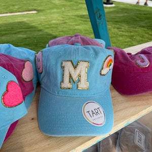 Może przedstawiać: Trzy czapki bejsbolowe w kolorze niebieskim, różowym i fioletowym z haftowanymi naszywkami. Niebieska czapka ma dużą białą literę "M" z brokatem złotym, chmurę tęczową i uśmiechniętą buźkę. Różowa czapka ma różowe serce, a fioletowa czapka ma fioletową literę "J". Wszystkie czapki mają białą naklejkę "TART DESIGN CO".