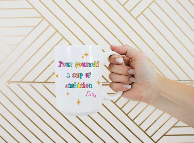 May include: White ceramic mug with a gold, pink, blue, and green design. The mug says "Pour yourself a cup of ambition - Dolly".