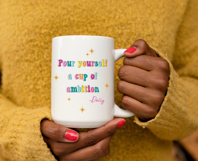 May include: White ceramic mug with a colorful quote that reads "Pour yourself a cup of ambition - Dolly". The quote is in a retro font with stars around it.