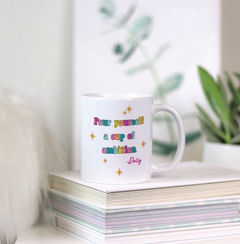 May include: White ceramic mug with a gold star design and the text "Pour yourself a cup of ambition - Dolly" in colorful letters.