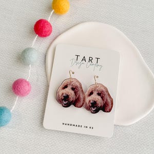 May include: A pair of gold dangle earrings featuring a brown dog's face on each earring. The earrings are displayed on a white card with the text "TART Design Company" and "HANDMADE IN KS".