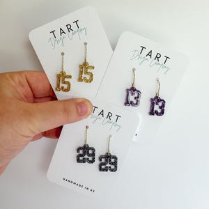 May include: Three pairs of glittery number earrings on white cards. One pair is gold with the number 15, another is purple with the number 13, and the third is black with the number 29. The cards read "TART Design Company" and "HANDMADE IN KS."