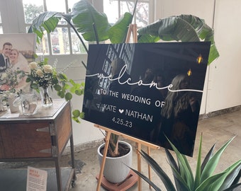 Puede incluir: Un cartel de bienvenida negro con texto blanco que dice "Bienvenido a la boda de Kate ♥ Nathan 4.25.23". El cartel está en un caballete y hay plantas en el fondo.