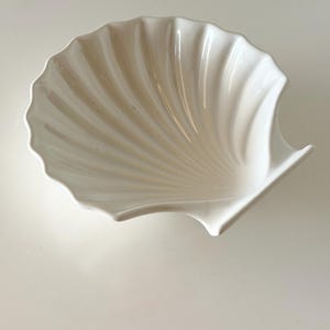 May include: A glossy white ceramic dish shaped like a scallop shell. The bowl features a smooth, curved interior with radiating ridges that extend from the base to the scalloped edge, creating a concave form with a clean, reflective finish.