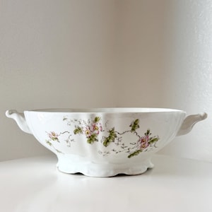 May include: A white ceramic serving bowl with a scalloped edge and two side handles. The exterior features a delicate botanical pattern of pink flowers, green leaves, and thin brown vines wrapping around the curved body of the vessel.