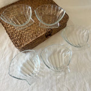 May include: Set of five clear glass seashell-shaped bowls. The bowls have a scalloped edge design and sit on a small, rectangular base. They are arranged on a woven wicker tray and a white fabric surface.