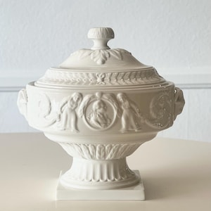 May include: White ceramic lidded serving dish with classical relief designs. The dish features a pedestal base and a decorative handle on the lid. The exterior is adorned with detailed figures and floral motifs, creating an elegant aesthetic.
