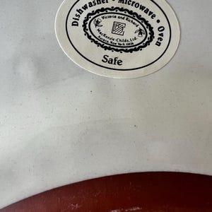 Pu&ograve; includere: Un piatto in ceramica bianco e rosso con un adesivo circolare. L'adesivo recita "Dishwasher Microwave Oven Safe" e presenta il testo "Victoria and Richard" e "Mackenzie-Childs. Ltd. Aurora, New York 13026."