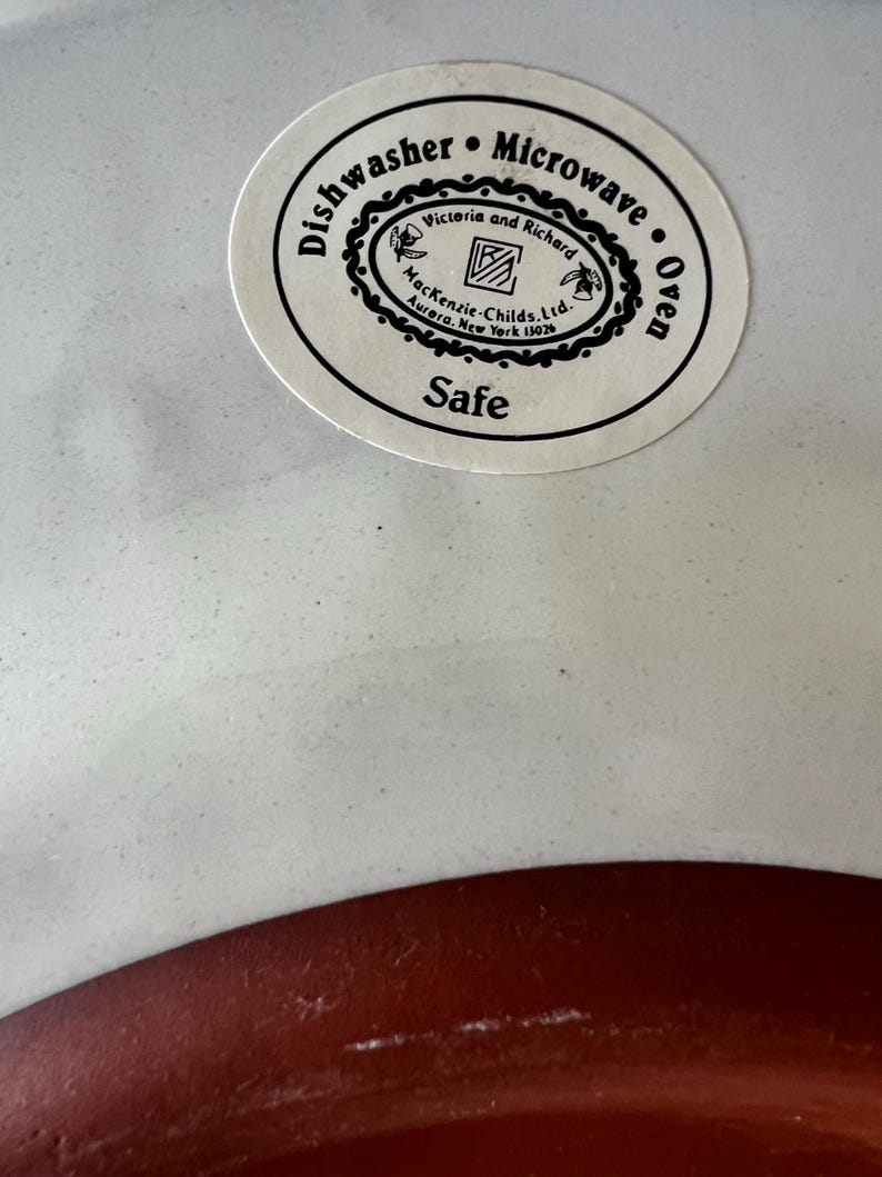 Pu&ograve; includere: Un piatto in ceramica bianco e rosso con un adesivo circolare. L'adesivo recita "Dishwasher Microwave Oven Safe" e presenta il testo "Victoria and Richard" e "Mackenzie-Childs. Ltd. Aurora, New York 13026."