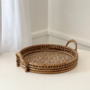 May include: A round, brown woven rattan tray featuring a flat base with a detailed herringbone pattern, a raised circular rim with a decorative zigzag weave, and two arched handles attached to opposite sides of the tray's outer edge.