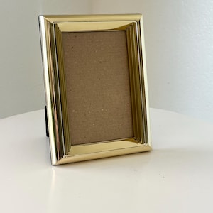 May include: A rectangular gold-colored picture frame with a ridged border. The frame has a black backing and a brown interior. The frame is standing upright on a white surface. The frame is designed to hold a photograph or artwork.