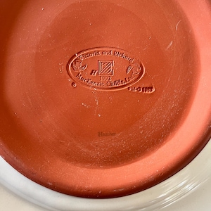 Pu&ograve; includere: Primo piano del fondo di un piatto in ceramica. Il piatto presenta un centro color terracotta con il testo in rilievo "Victoria and Richard Mackenzie-Childs, Ltd. 1992" e la parola "Heather". Il bordo esterno &egrave; bianco.