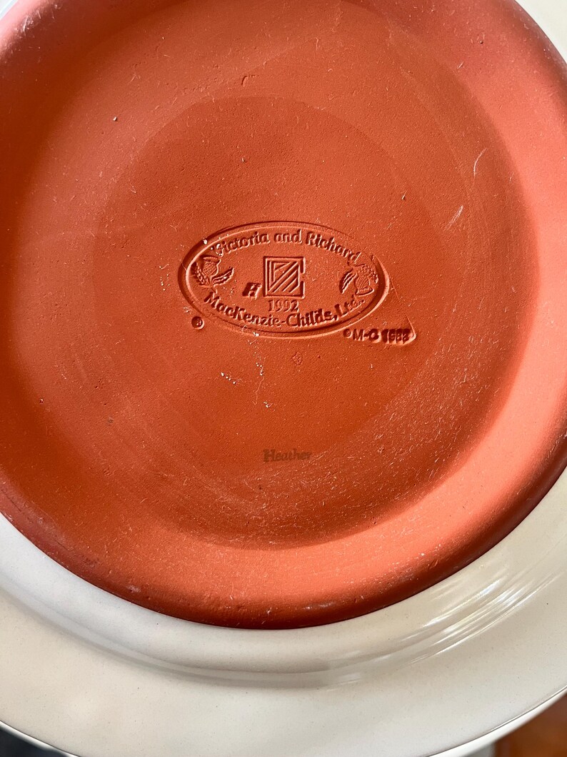 Pu&ograve; includere: Primo piano del fondo di un piatto in ceramica. Il piatto presenta un centro color terracotta con il testo in rilievo "Victoria and Richard Mackenzie-Childs, Ltd. 1992" e la parola "Heather". Il bordo esterno &egrave; bianco.
