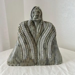 May include: A gray stone bust of a Native American figure, wearing a hooded cloak with feather and fringe details. The sculpture has a solemn expression and is set against a white background.