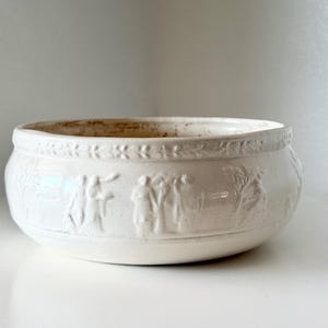 May include: A round, off-white ceramic bowl featuring a raised relief design around the exterior. The pattern includes a band of repeating leaf motifs near the rim and a series of figures and trees embossed along the center of the vessel wall.