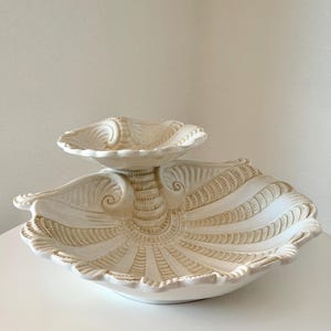 May include: A two-tiered ceramic serving dish featuring a shell-inspired design. The piece is finished in an off-white glaze with light brown detailing highlighting the textured, radiating ridges and scalloped edges of the two bowl sections.