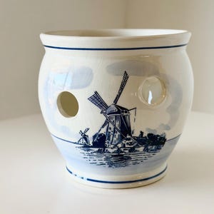 May include: A white ceramic pot with a blue windmill design. The pot has two circular holes and a blue rim. The windmill scene depicts a Dutch landscape with windmills and water. The pot is likely for kitchen or home decor.