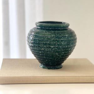 May include: A dark teal ceramic vase with a textured, ribbed design. The vase has a rounded body, a narrow neck, and a small base. It sits on a beige book with the title "Masterpieces of Tutankhamun".