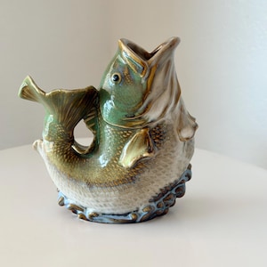 May include: A ceramic fish-shaped pitcher with a glazed finish in shades of green, brown, and cream. The pitcher features a wide-open mouth at the top, a curved tail handle, and textured scales along the body, with a dark blue base detail.