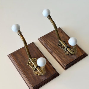 May include: Two wall-mounted coat hooks, each featuring a rectangular dark wood base, a curved brass metal arm, and two white ceramic ball-shaped knobs at the ends. The brass hardware is secured to the wood with visible screws at the base.