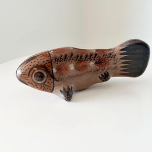 May include: A hand-painted ceramic fish figurine in shades of brown, tan, and black. The body features intricate dark brown line work, small dots, and feather-like patterns along the dorsal area, with a solid black tail and small fins.