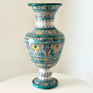 May include: A tall, decorative ceramic vase with a pedestal base. The vase features a vibrant design with teal, yellow, and blue geometric patterns. The rim and base are teal, and the body has intricate floral motifs. The vase is approximately 12 inches tall.