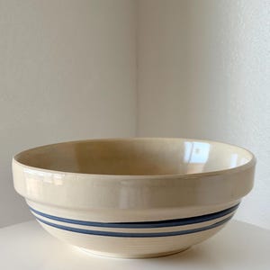 May include: A cream-colored ceramic mixing bowl with a smooth, glossy finish. The exterior features two distinct, horizontal dark blue bands encircling the lower portion of the bowl. The bowl has a wide, open top and a slightly rounded base.