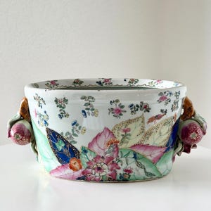 May include: A white ceramic bowl featuring colorful hand-painted floral and leaf patterns in shades of pink, green, and blue. The bowl has two decorative handles shaped like fruit, and the interior rim is decorated with small floral motifs.