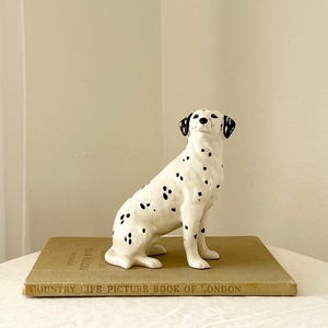 May include: A ceramic Dalmatian dog figurine with black spots, sitting atop a vintage book titled "COUNTRY LIFE PICTURE BOOK OF LONDON." The figurine is white with black spots and black ears. The book is a light brown color.