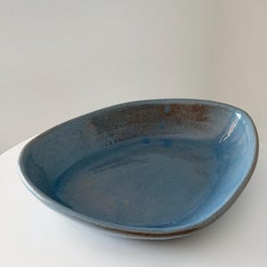 May include: A shallow, asymmetrical ceramic bowl with a glossy blue glaze. The interior features a smooth, reflective surface with subtle variations in tone, while the outer rim displays a textured, brownish-grey finish along the edge.