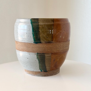 May include: A ceramic planter with a rounded shape featuring a speckled light gray glaze on the left, a smooth brown glaze on the right, and a vertical dark green stripe. A textured, unglazed horizontal band wraps around the center.