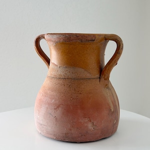 May include: A rustic terracotta vessel featuring a wide, flared rim and two symmetrical side handles. The upper portion is finished in a smooth, ochre-toned glaze, while the lower body displays a textured, matte reddish-brown clay surface.