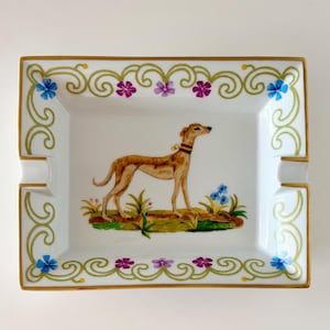 May include: A rectangular white porcelain ashtray featuring a hand-painted tan greyhound wearing a red collar. The border is decorated with green scrollwork, blue and purple flowers, and a gold-painted rim with two cigarette rests.