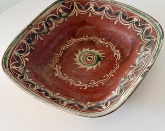 May include: A square terracotta bowl featuring a hand-painted sun motif in the center surrounded by a circular border. The rim is decorated with a repeating wavy line pattern in green, dark blue, and cream, set against a deep reddish-brown base.